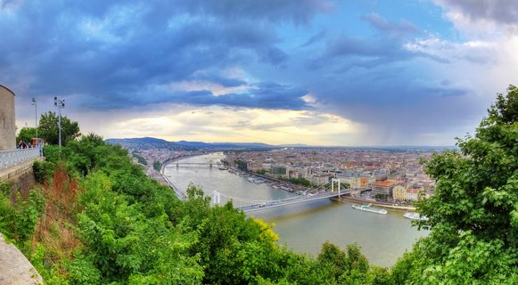 Panoramic cityscape of Budapest showing the Chain Bridge crossing the Danube River with dramatic cloudy sky and green vegetation in foreground