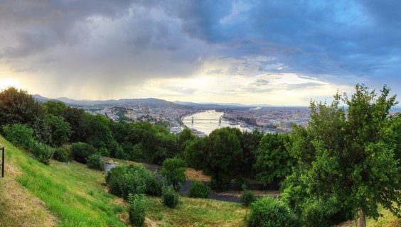 Panoramic cityscape of Budapest showing the Danube River with Chain Bridge, Buda Castle on the hillside, and dramatic cloudy sky with sun rays during sunset, viewed from an elevated position with green trees in foreground