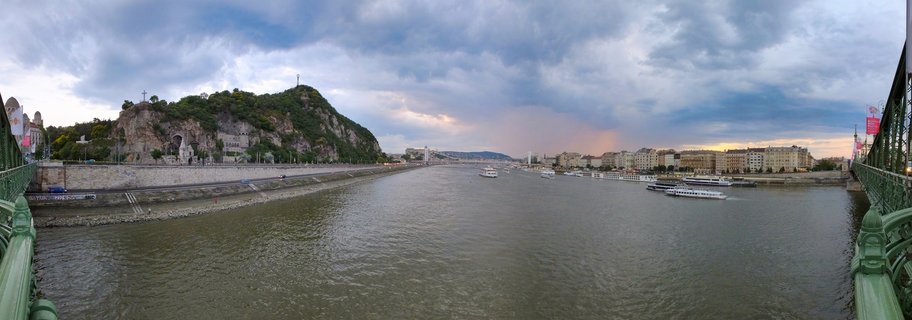 Wide panoramic photograph of Budapest's Danube River showing Gellért Hill with Liberty Statue on the left, multiple white cruise boats on the water, and historic buildings along the right riverbank, viewed from Liberty Bridge with green metal bridge structure visible at edges
