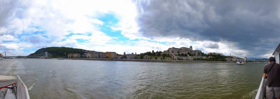 Wide panoramic photograph showing Budapest's Buda Castle on a hill overlooking the Danube River, with the Chain Bridge visible on the left side and dramatic cloudy sky above