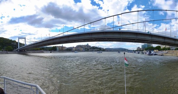 White suspension bridge spanning wide river with castle on hillside in background, Hungarian flag visible in foreground, boats on water