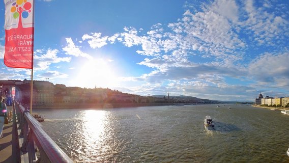 Panoramic photograph of Budapest showing the Danube River with the Hungarian Parliament Building on the right, Buda Castle on the left hillside, tourist boats on the water, and a red festival banner in the foreground during golden hour sunlight