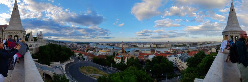 Panoramic view from Fisherman's Bastion showing stone turrets, tourists on terraces, Danube River, and Budapest cityscape with Parliament Building under blue sky with clouds