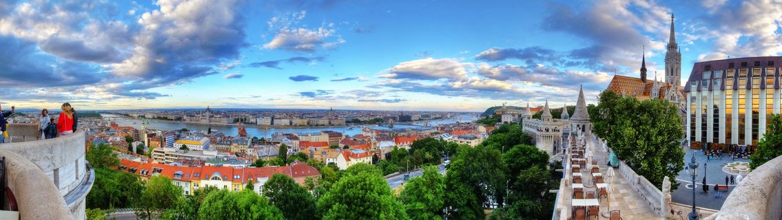 Wide panoramic view of Budapest cityscape from Fisherman's Bastion showing the Danube River, red-roofed buildings, Matthias Church spire, and Hungarian Parliament in the distance under a cloudy blue sky