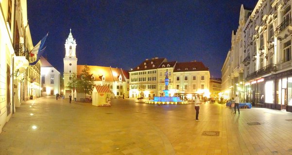 Night view of Bratislava Main Square showing illuminated Old Town Hall tower, blue-lit Maximilian Fountain, historic buildings, and pedestrians on cobblestone plaza