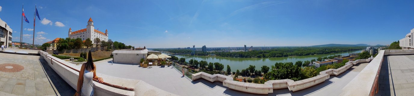 Panoramic cityscape showing Bratislava Castle with white walls and red towers on a hill, the Danube River flowing through green landscape, and modern city buildings under a clear blue sky
