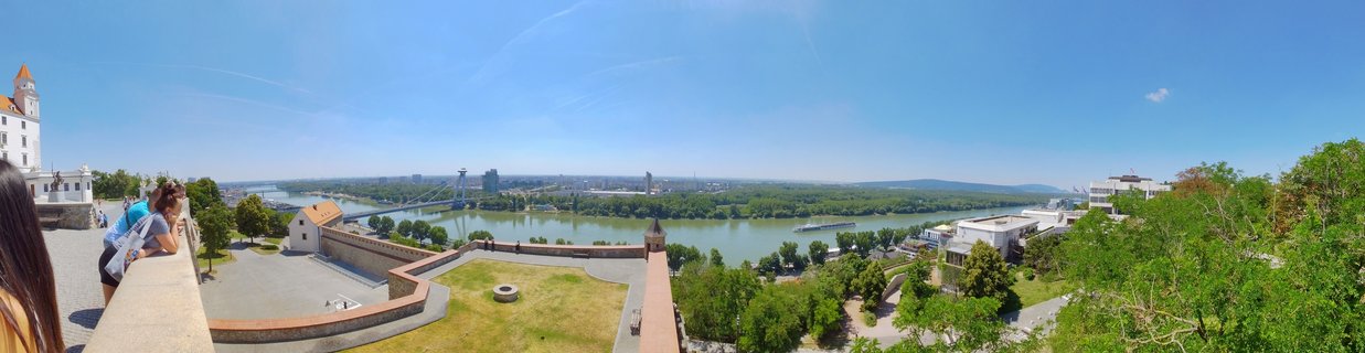 Panoramic cityscape showing Bratislava Castle's white walls and red tower on the left, the wide Danube River in the center with UFO Bridge spanning across, and green trees along the riverbanks under clear blue sky