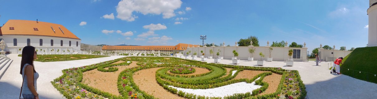Panoramic view of formal baroque gardens at Bratislava Castle with geometric hedge designs, white buildings with orange roofs, and visitors walking on gravel paths under blue sky