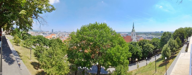 Panoramic cityscape of Bratislava showing St. Martin's Cathedral spire, red-roofed Old Town buildings, green trees, and blue sky