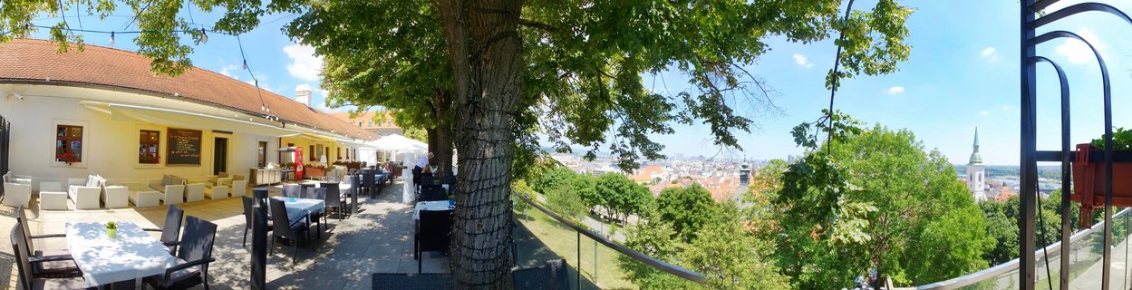 Outdoor restaurant terrace with tables and chairs under trees, overlooking Bratislava city with church towers visible in the distance on a sunny day