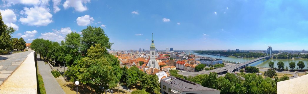Panoramic cityscape of Bratislava showing St. Martin's Cathedral tower, red-roofed Old Town buildings, green trees, the Danube River, and the UFO Bridge under blue sky with white clouds