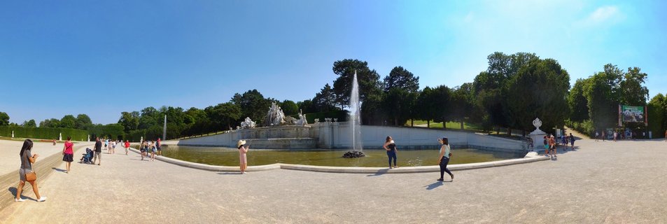 Wide panoramic view of the Neptune Fountain at Schönbrunn Palace with baroque statues, water fountains, gravel pathways, and tourists walking in the palace gardens under a clear blue sky