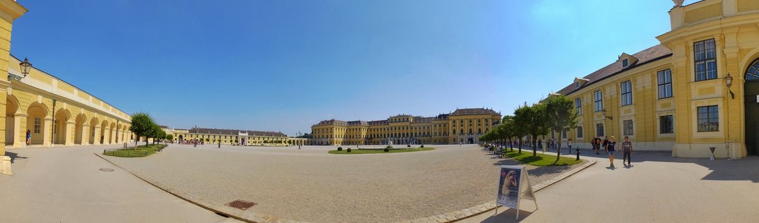 Wide panoramic view of Schönbrunn Palace in Vienna showing the yellow baroque main building in the distance, flanked by arched colonnade wings on both sides, with tourists walking across the gravel courtyard under a clear blue sky