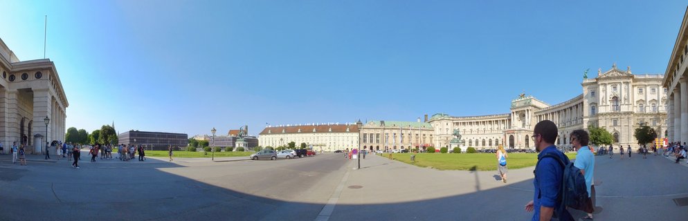 Wide panoramic view of Heldenplatz square in Vienna showing the Hofburg Palace buildings with tourists walking on paved areas and green lawns under clear blue sky