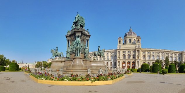 Large bronze equestrian monument with multiple statues on ornate pedestal, grand baroque museum building with dome in background, blue sky, flower beds in foreground