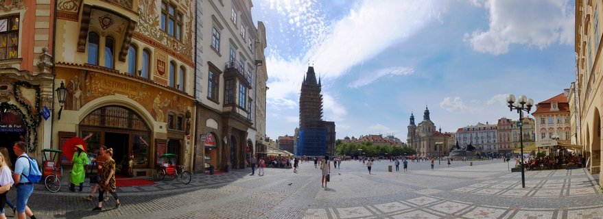 Wide view of Prague's Old Town Square showing historic buildings with ornate facades, tourists walking on cobblestone pavement, and the Old Town Hall Tower covered in scaffolding against a blue sky with clouds