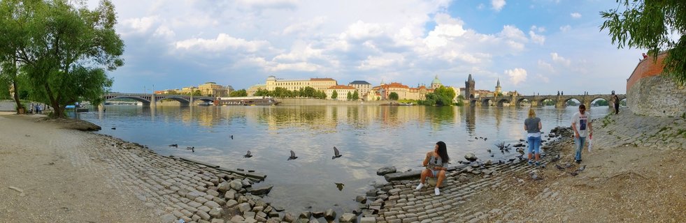 Wide panoramic photograph of Prague showing the Vltava River with Charles Bridge on the right, historic buildings along the far bank, people sitting on the cobblestone shore, and trees framing the scene under a partly cloudy sky