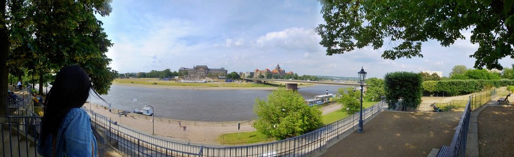 Panoramic view of Dresden's historic buildings across the Elbe River with a woman in denim jacket looking at the skyline from an elevated terrace with metal railings and trees