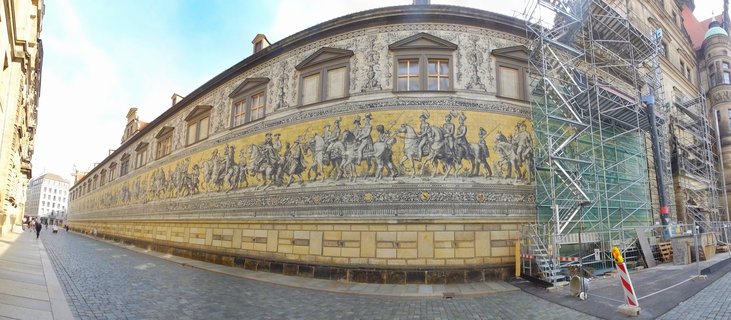 A long panoramic view of the Fürstenzug mural showing a procession of mounted Saxon rulers and nobles on a building wall along a cobblestone street in Dresden, with scaffolding covering part of the right side.