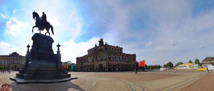 Panoramic view of the Semperoper opera house in Dresden with the King Johann equestrian statue on cobblestone Theaterplatz under blue sky with clouds