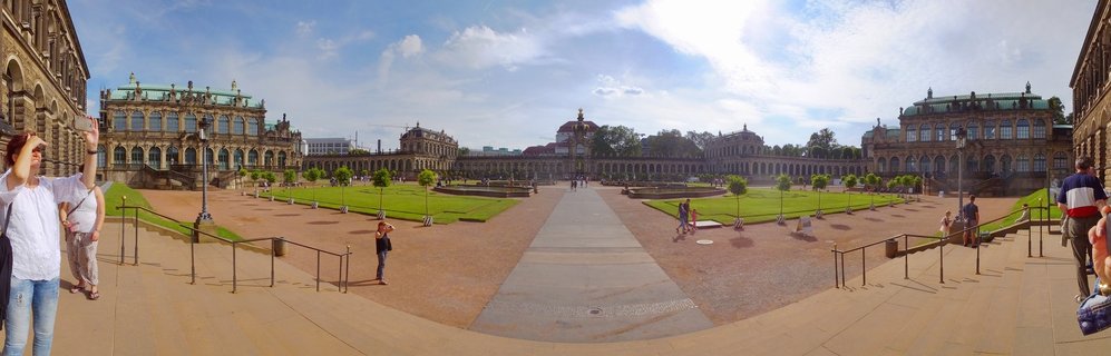 Panoramic view of the Zwinger Palace courtyard in Dresden showing baroque buildings with green copper roofs, manicured lawns, stone pathways, and tourists visiting the historic landmark under a blue sky with scattered clouds