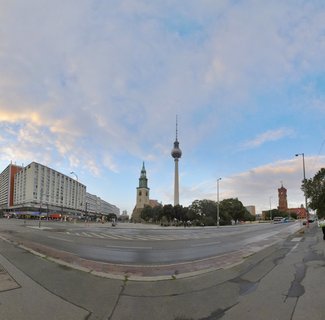 Panoramic street view of Berlin showing the TV tower, church with green spire, red city hall building, wide intersection, and cloudy evening sky