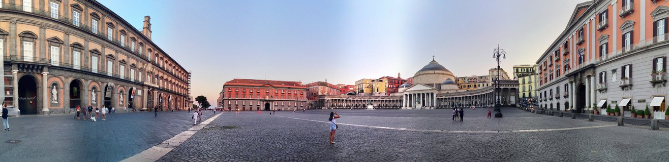 Wide panoramic view of Piazza del Plebiscito in Naples showing the Royal Palace on the left, the domed Church of San Francesco di Paola with colonnades on the right, and people walking across the cobblestone square at sunset