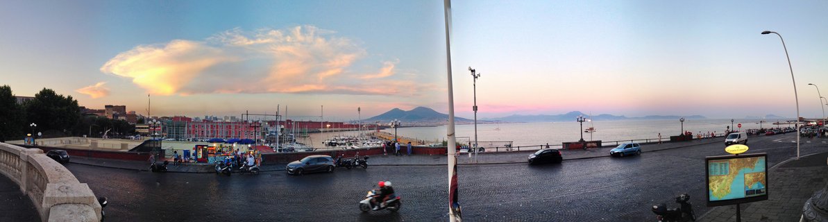 Panoramic street view of Naples waterfront at sunset showing Mount Vesuvius across the bay, with cars, motorcycles, pedestrians, marina boats, and colorful sky