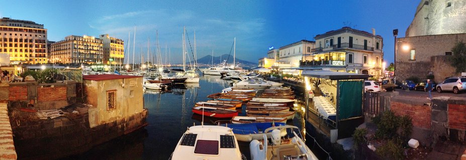 Panoramic harbor scene showing multiple boats docked in a marina with illuminated buildings along the waterfront and mountains visible in the background during twilight