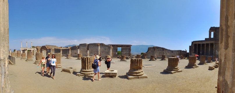 Ancient Roman stone columns and ruins at Pompeii archaeological site with tourists walking among the structures and Mount Vesuvius visible in the background under clear blue sky