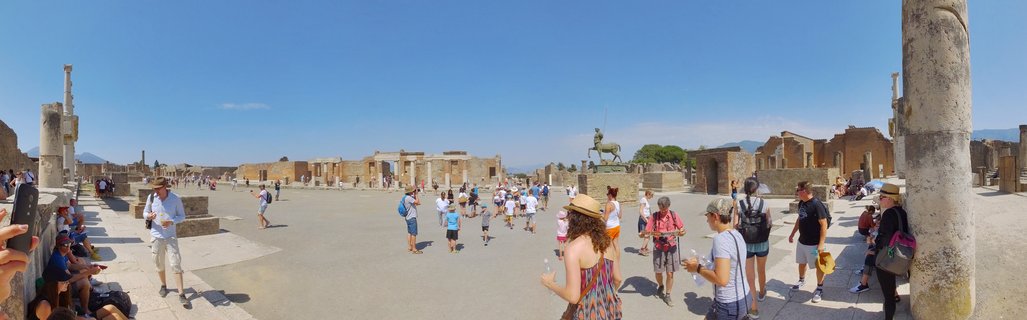 Wide panoramic view of tourists walking among ancient Roman stone ruins and columns at Pompeii archaeological site under clear blue sky with Mount Vesuvius visible in distance