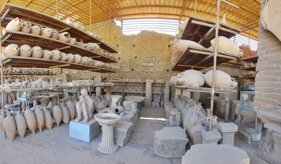 Indoor archaeological storage showing rows of ancient Roman pottery, amphorae on metal shelving, stone column fragments, and a plaster cast human figure at Pompeii ruins