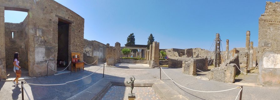 Ancient Roman ruins at Pompeii archaeological site showing stone courtyard with weathered walls, columns, bronze statue in foreground, and visitor walking on left side under clear blue sky