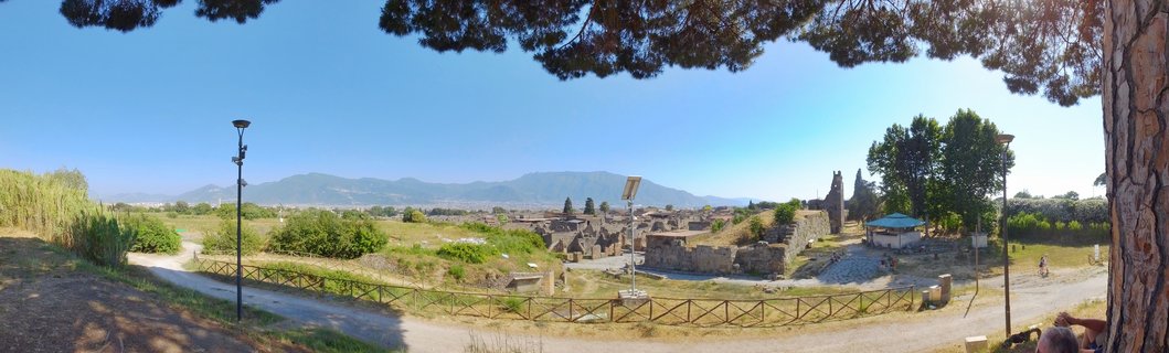 Wide panoramic view of ancient stone ruins at Pompeii archaeological site with Mount Vesuvius mountain range in background under clear blue sky, pine tree branches framing the top edge