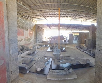 Multiple plaster casts of human bodies displayed on black platforms inside an archaeological exhibition hall with stone walls and protective roof structure
