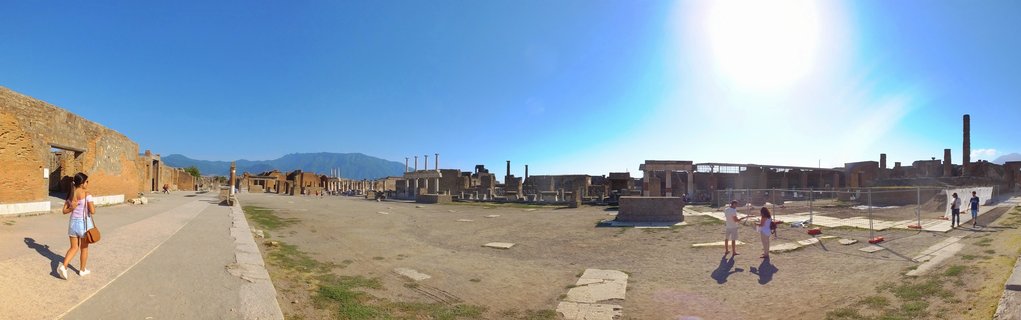 Panoramic view of ancient Roman ruins at Pompeii archaeological site showing stone columns, brick walls, and pathways with tourists walking under clear blue sky with Mount Vesuvius in background