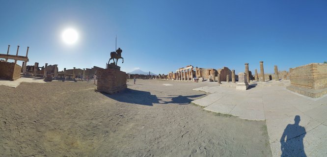Wide view of ancient Roman ruins at Pompeii's Forum with stone columns, brick walls, an equestrian statue, and Mount Vesuvius visible in the distance under clear blue sky
