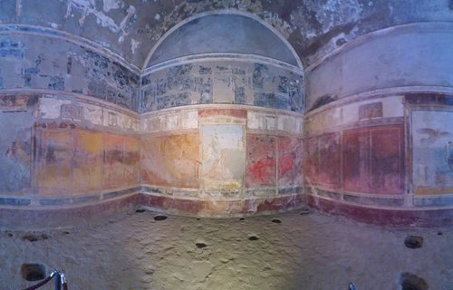 Interior of ancient Roman room in Pompeii showing faded wall frescoes with red yellow and blue painted panels on curved walls with stone floor
