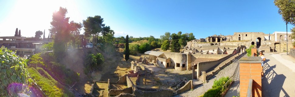 Panoramic view of ancient Roman stone ruins at Pompeii archaeological site with visitors, Mediterranean trees, and clear blue sky