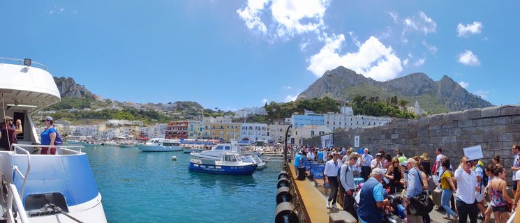 Panoramic view of a busy harbor with tourists on a dock, blue and white boats in turquoise water, colorful Mediterranean buildings on a hillside, and mountains under a blue sky with scattered clouds
