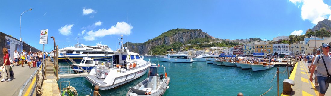 Panoramic view of Marina Grande harbor in Capri showing ferries docked in turquoise water, tourists walking on piers, colorful buildings on hillside, and limestone cliffs under blue sky