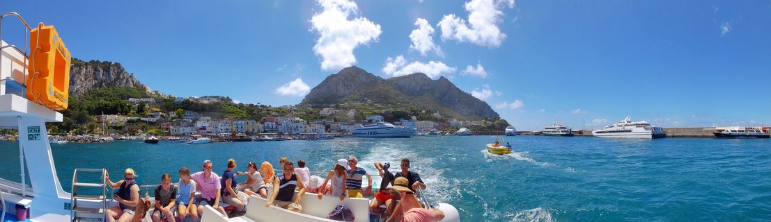 Tourists seated on a white boat in turquoise Mediterranean water approaching Capri's Marina Grande harbor with white buildings on hillsides, large ferries, and limestone cliffs under blue sky with clouds