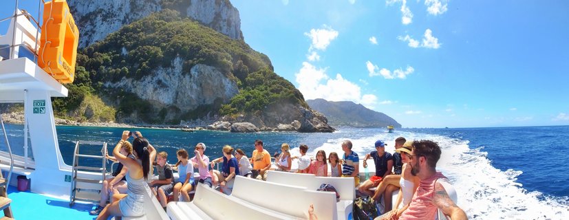 Group of tourists sitting on a white boat deck with orange life preserver, cruising on blue Mediterranean Sea with green-covered limestone cliffs of Capri in background under partly cloudy sky