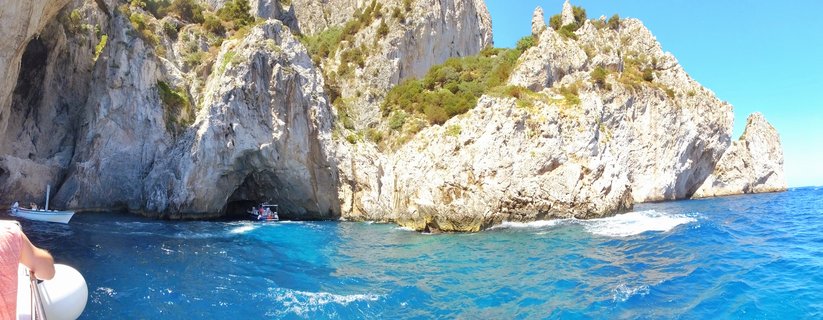 White limestone cliffs with sea cave entrance rising from turquoise Mediterranean water near Capri, Italy, with tourist boats approaching the grotto under clear blue sky