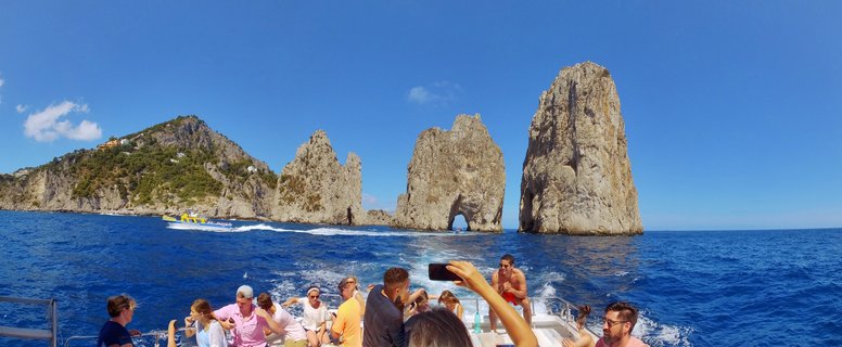 Tourists on a boat taking photos of three large rock formations rising from blue Mediterranean water near Capri, Italy under a clear blue sky