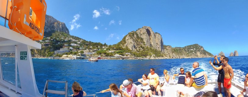 Tourists on a white ferry boat deck viewing Capri island coastline with steep limestone cliffs, white buildings on hillsides, and Faraglioni rock formations in the blue Mediterranean Sea under clear sky