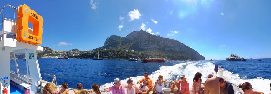 Tourists on a ferry deck with orange life preserver visible, approaching Capri island with mountain cliffs under blue sky with scattered clouds