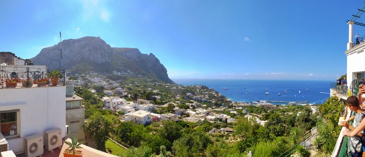Wide panoramic photograph showing Capri island's white buildings on green hillsides, dramatic mountain cliffs, and blue sea with boats under clear sky
