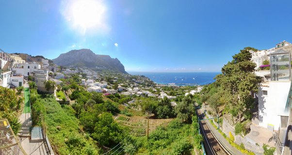 Panoramic landscape showing Capri island in Italy with white buildings on green hillsides, funicular railway tracks, Mount Solaro mountain in background, and blue Mediterranean sea with boats