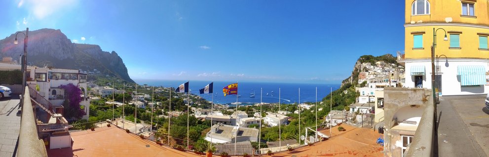 Panoramic photograph of Capri, Italy showing white buildings on hillsides, blue Mediterranean Sea with boats, yellow building on right, and limestone mountain cliffs in the distance under clear sky